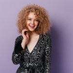 Pretty girl with blond curls cute smiles. Snapshot of lady in shiny v-neck blouse on purple background.